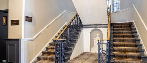 A grand staircase with a checkered carpet and black balusters, leading to a landing with a statue.