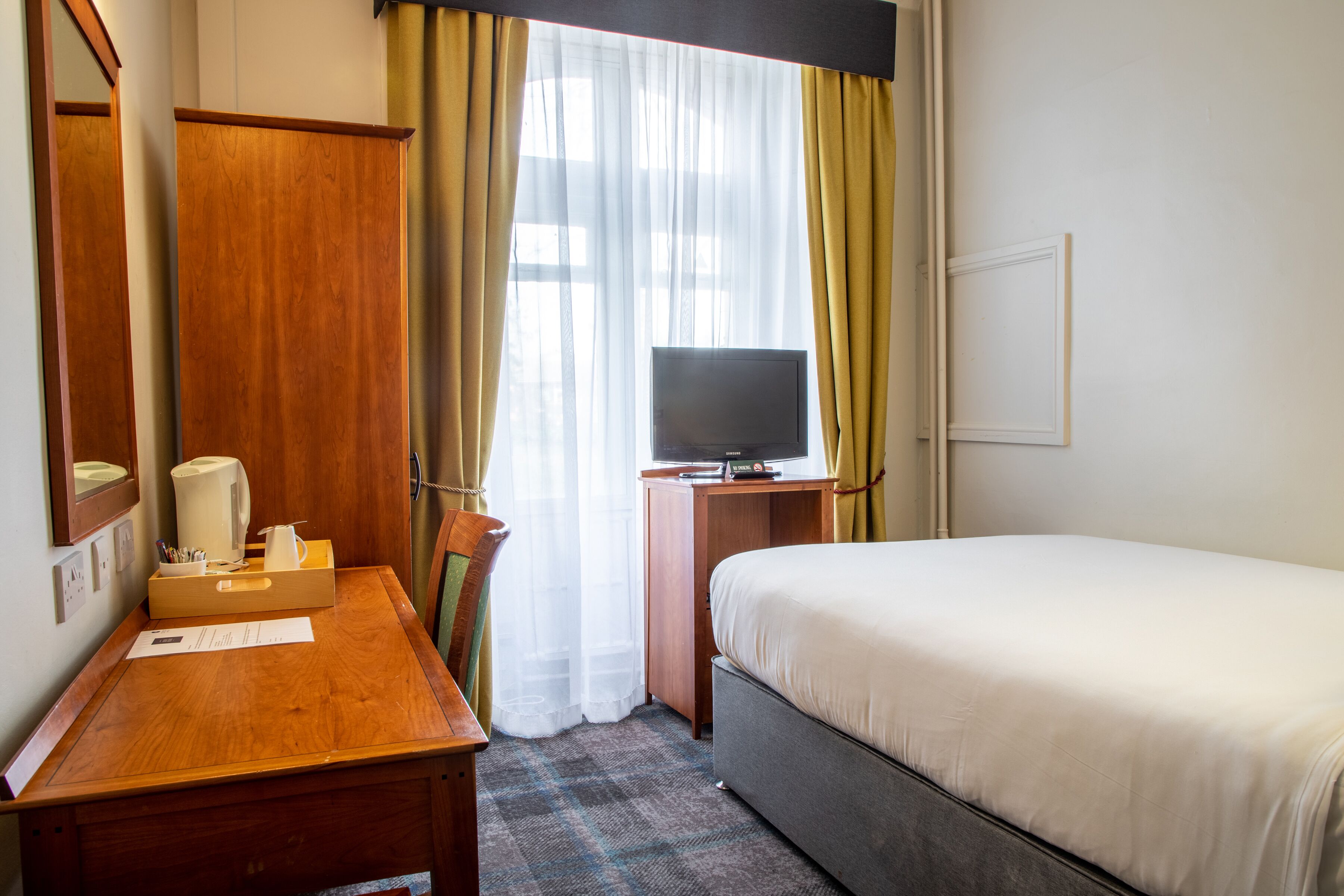 A hotel room with a bed, a desk with a mirror, a television, and a window with curtains.