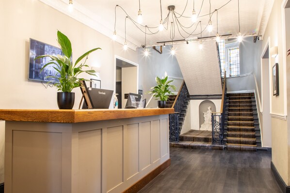 A reception area with a wooden counter, potted plants, a desk with a computer, and a staircase leading to an upper floor. - Arnos Manor Hotel (Bristol)