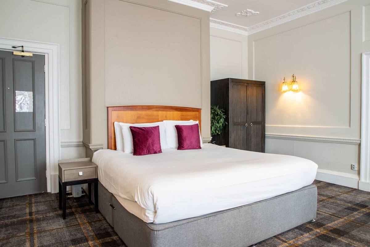 Una habitación de hotel ordenada con una cama grande, una mesita de noche, un armario y una lámpara fijada a la pared.