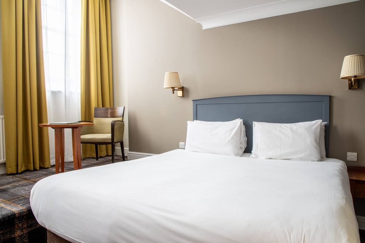 Habitación de hotel con una cama grande, una mesita, una silla y dos lámparas fijadas a la pared.