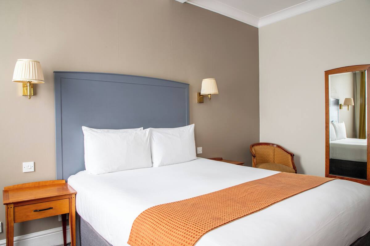 Una cama bien tendida con sábanas blancas, cabecera azul y una manta naranja. Hay una mesita de noche de madera con un cajón y una lámpara encima. Una silla de mimbre está colocada cerca de la cama, y hay un espejo en la pared.