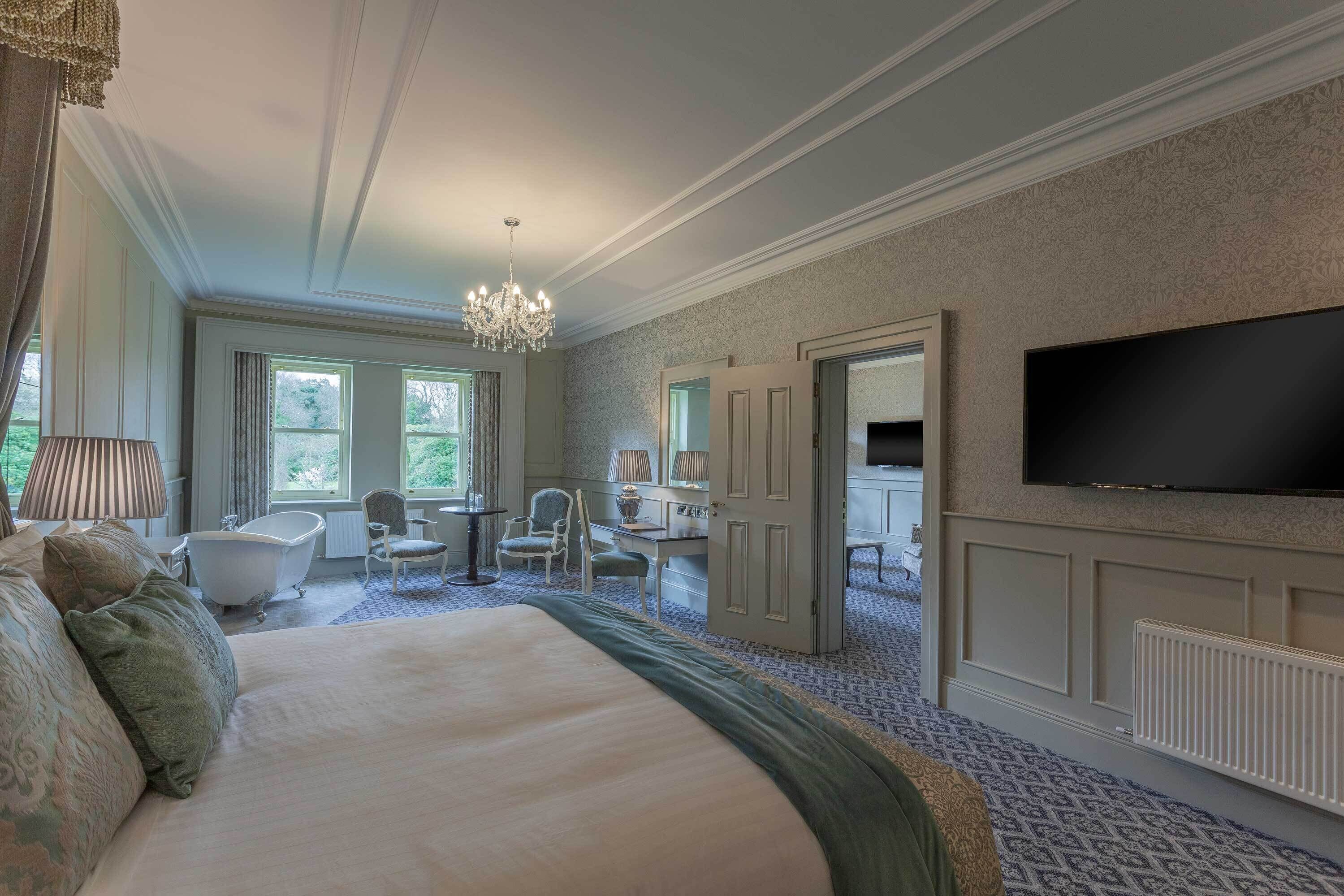 A spacious bedroom with a large bed, a sitting area with a chair and a table, a bathtub, and a television mounted on the wall.