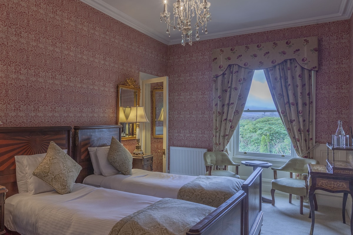 A hotel room with two beds, a sitting area with a table and chairs, a chandelier, and a view through the window.