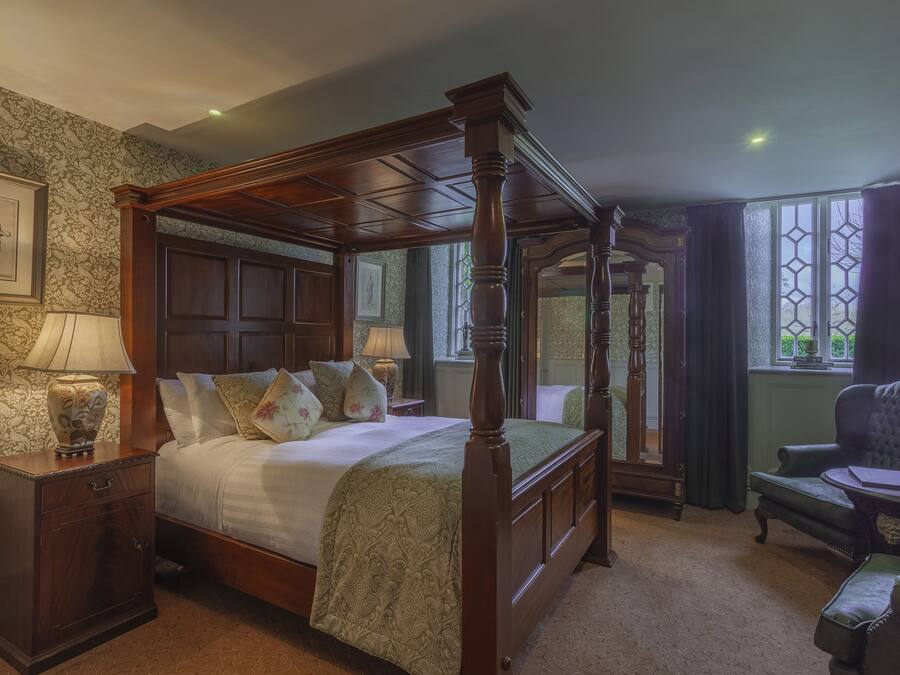 A four-poster bed with a wooden frame, a nightstand with a lamp, a chair, and a window with curtains.