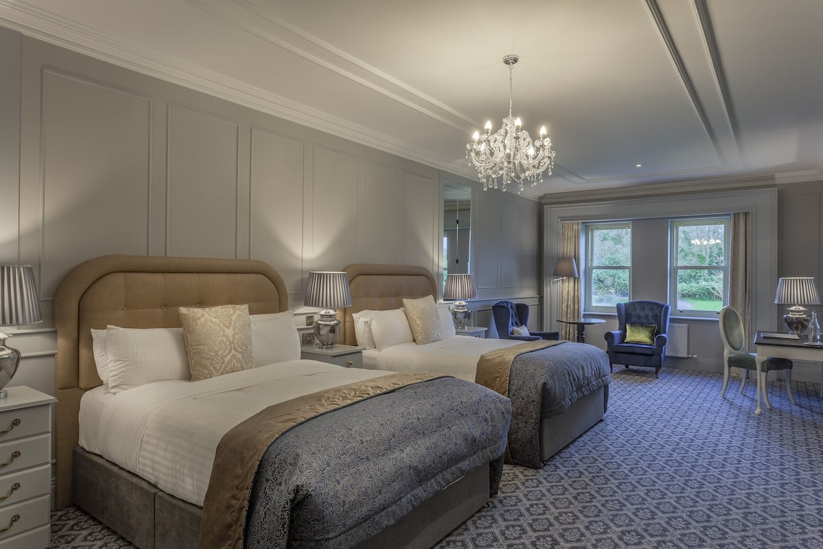 A hotel room with two beds, a chandelier, a desk, and a view of the outdoors.
