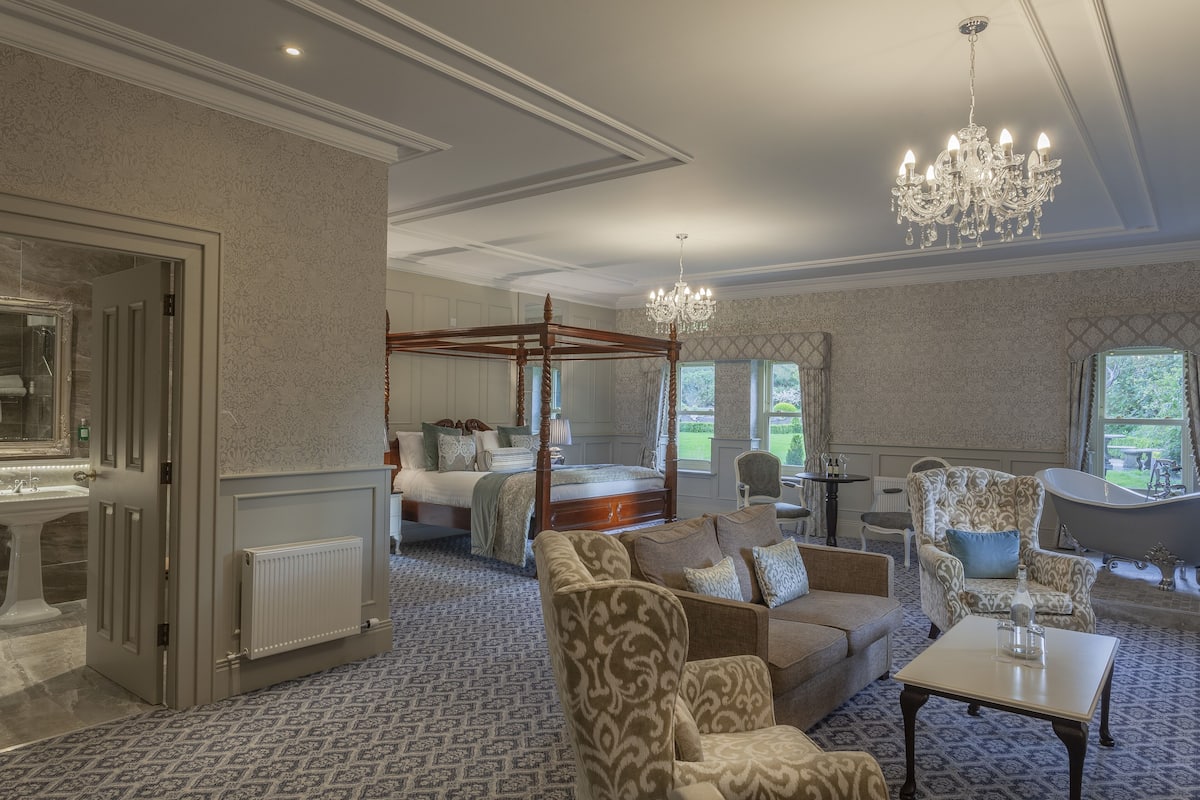 A spacious bedroom with a four-poster bed, a sitting area with patterned armchairs, a large chandelier, and a view of the garden through the windows.
