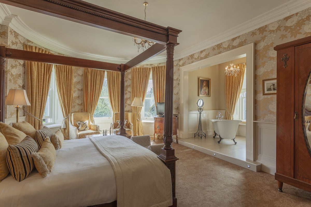 A spacious bedroom with a four-poster bed, a sitting area with a chair and table, and a view of the bathroom.