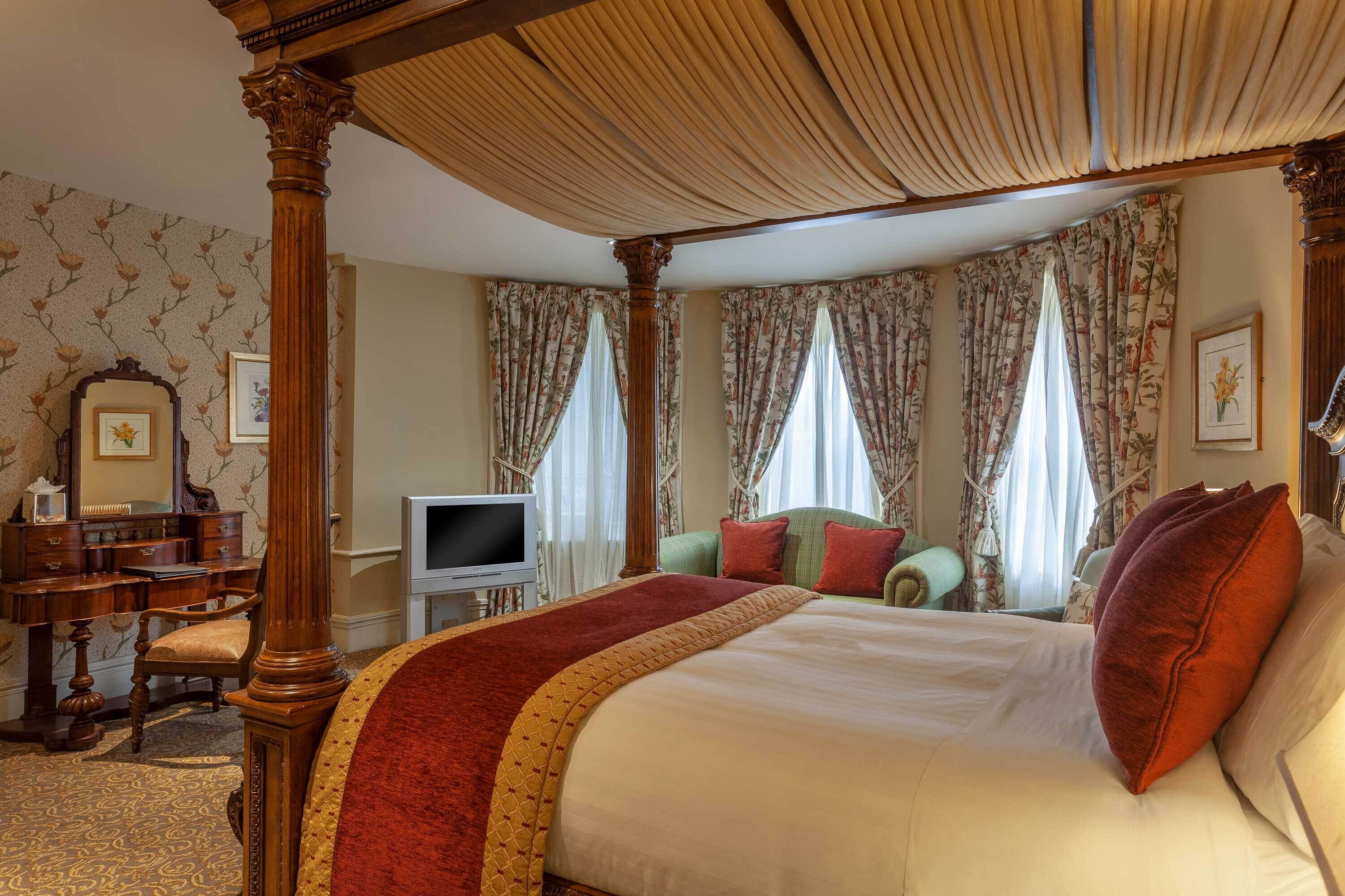 Una cama con dosel adornada con una colcha roja y dorada, un televisor y un escritorio de madera con una silla en una habitación con papel tapiz y cortinas florales.