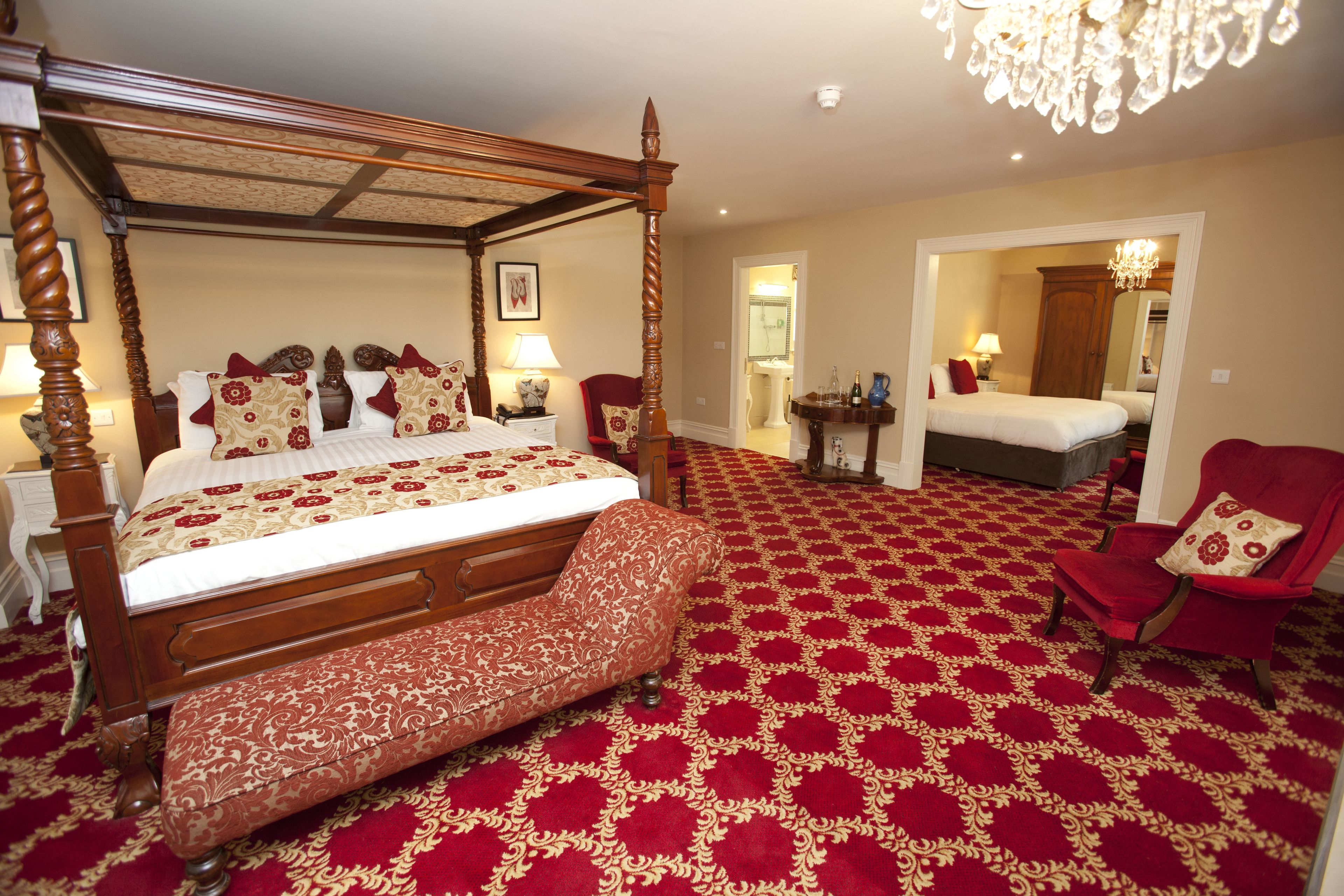 A four-poster bed with a red and gold patterned bedspread, a wooden bench, and a red armchair in a room with a red and gold carpet.