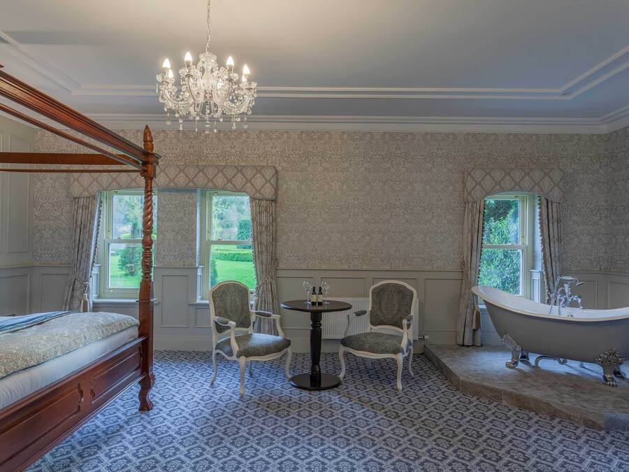 A four-poster bed, two upholstered chairs, a round table with a vase, and a freestanding bathtub in a room with patterned wallpaper and large windows.