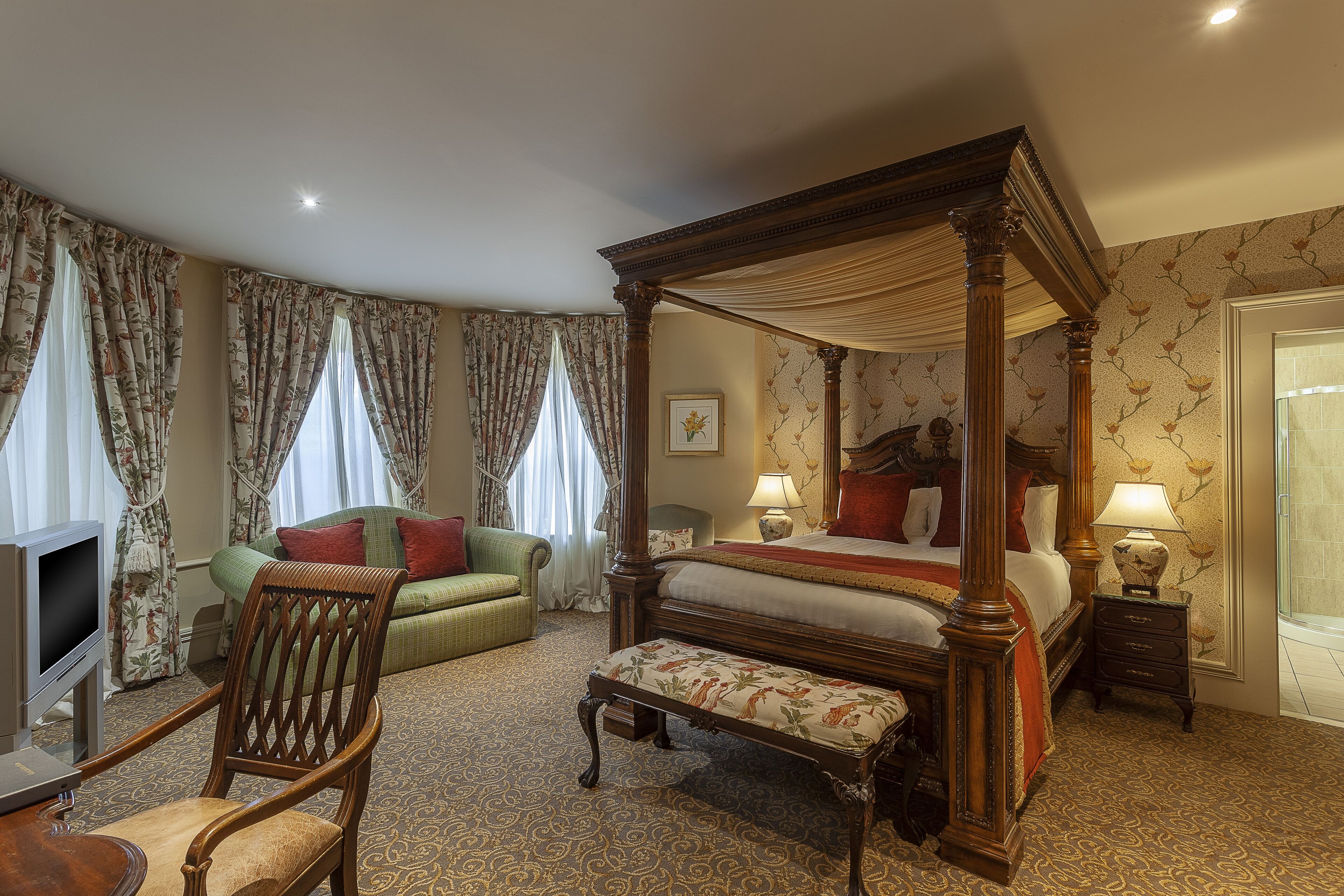 A four-poster bed with a wooden frame, a green upholstered sofa, a wooden chair, a small table, and a television.