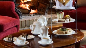 A dining table with a three-tiered serving tray, a teapot, cups, and plates with cutlery. A red armchair is visible to the left, and a fireplace with a fire is in the background. - Buckland Manor (Broadway)