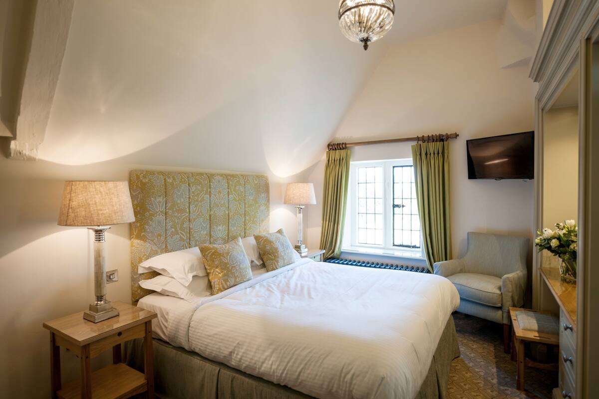 Une chambre bien rangée, avec un lit, deux tables de chevet, une lampe, une chaise, un téléviseur et une fenêtre avec des rideaux.