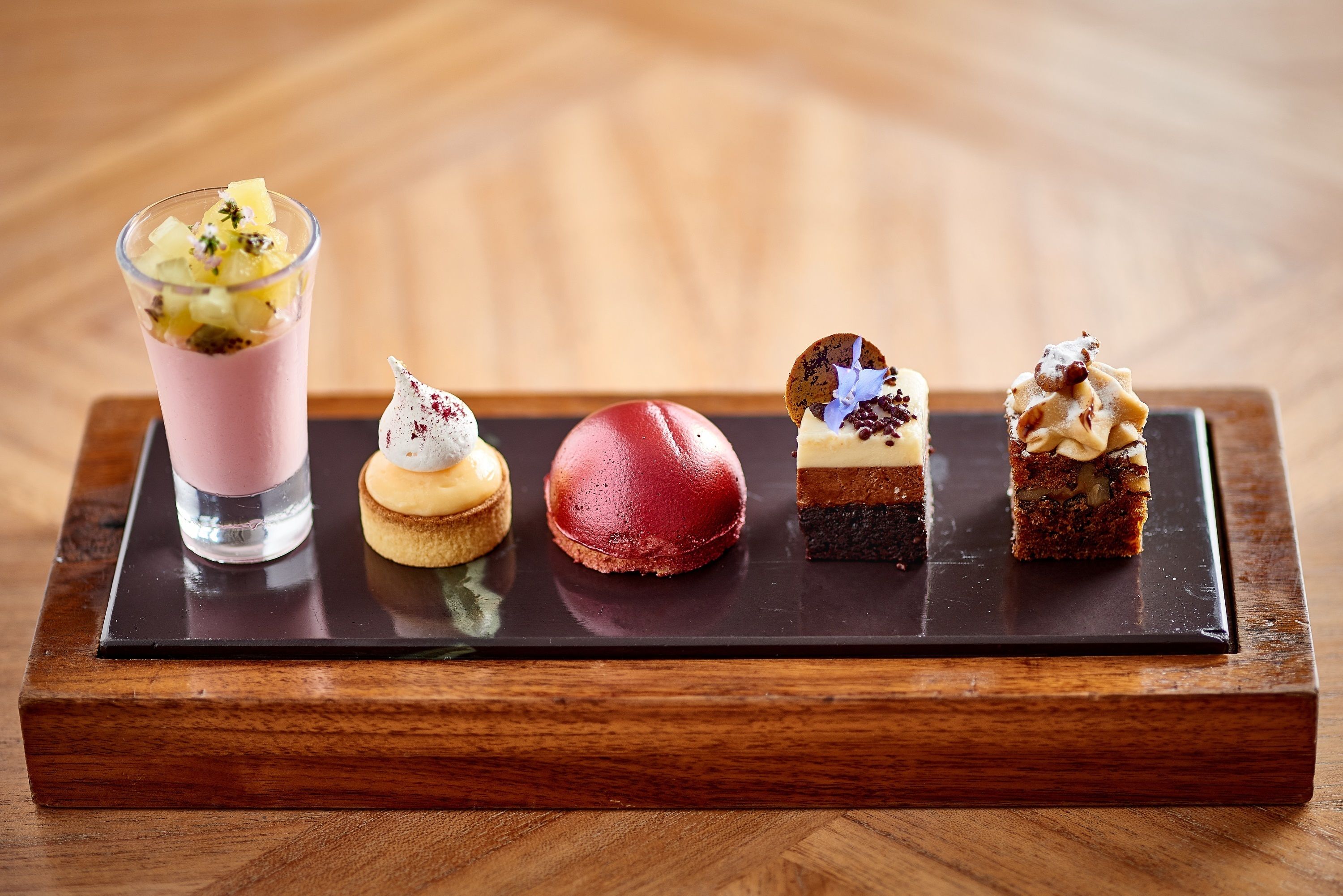 Una bandeja de postres con diversas golosinas, incluyendo una bebida rosa, un macaron rojo, un profiterole blanco, un postre de chocolate y un pastel en capas.