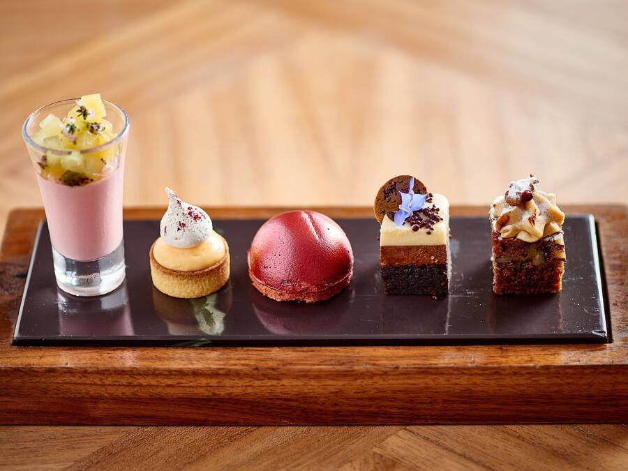 Una bandeja de postres con diversas golosinas, incluyendo una bebida rosa, un macaron rojo, un profiterole blanco, un postre de chocolate y un pastel en capas.