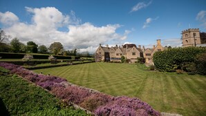 A large, historic building with a symmetrical facade, surrounded by well-manicured lawns and a variety of flowering plants. - Buckland Manor (Broadway)
