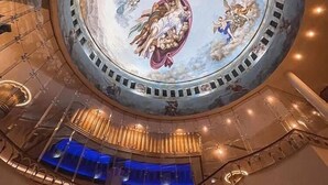 A grand atrium with a circular ceiling mural depicting classical figures and scenes, surrounded by a modern glass and metal structure.