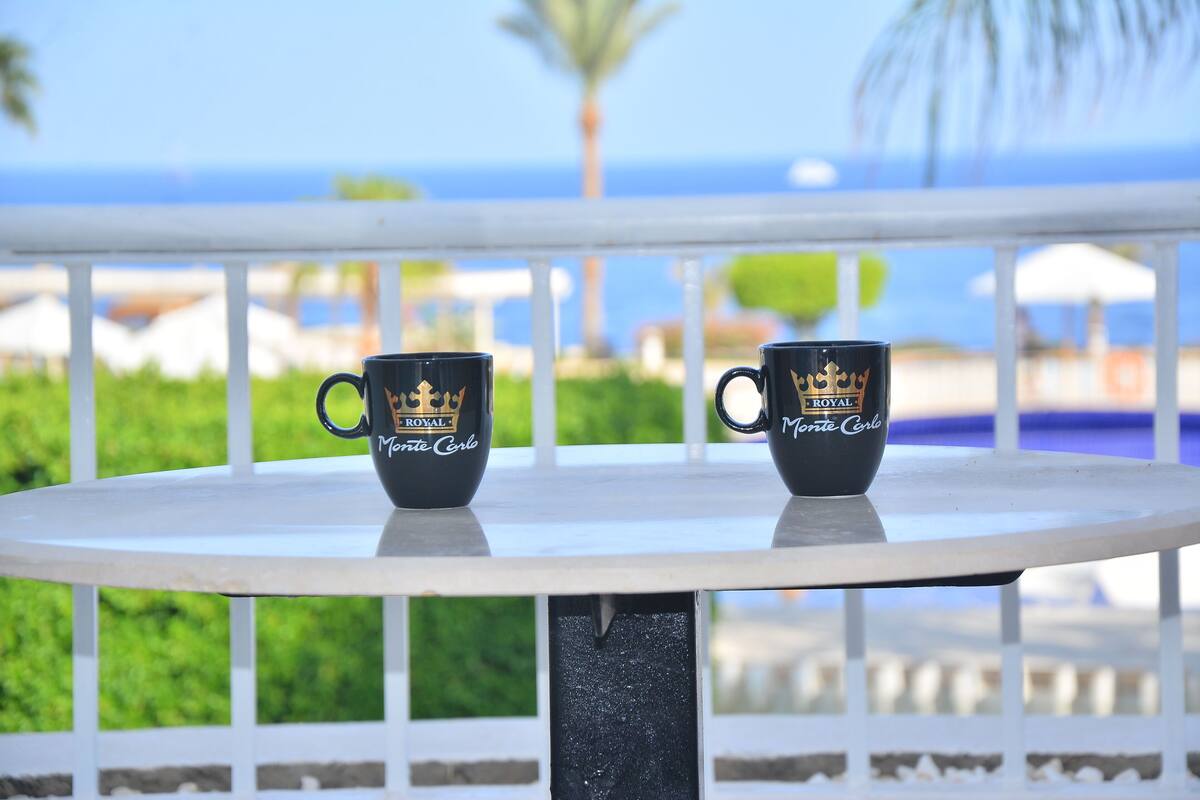 Two black mugs with gold text on a white table outdoors with a view of the ocean and palm trees.
