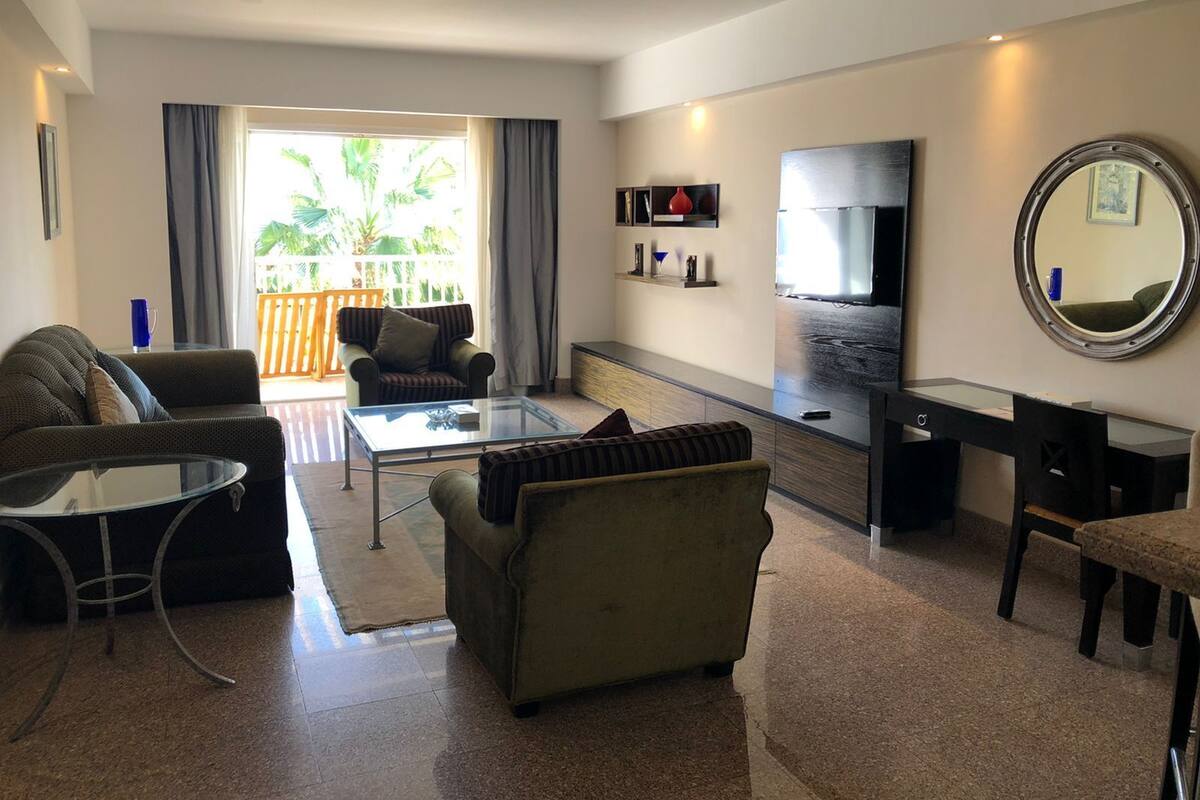A modern living room with a sofa, armchair, and coffee table. A flat-screen TV and a round mirror are mounted on the wall.