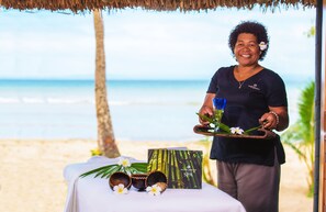 A person standing at a table with a beach view, holding a tray with a drink and a small plant. There are spa-related items on the table, including a card and a small bowl.