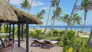 Une terrasse en bois avec des chaises longues et une structure à toit de chaume, donnant sur une plage bordée de palmiers et sur l’océan.