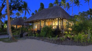 Un bâtiment à toit de chaume, doté d’une terrasse en bois et de sièges extérieurs, entouré d’une végétation tropicale et de palmiers.