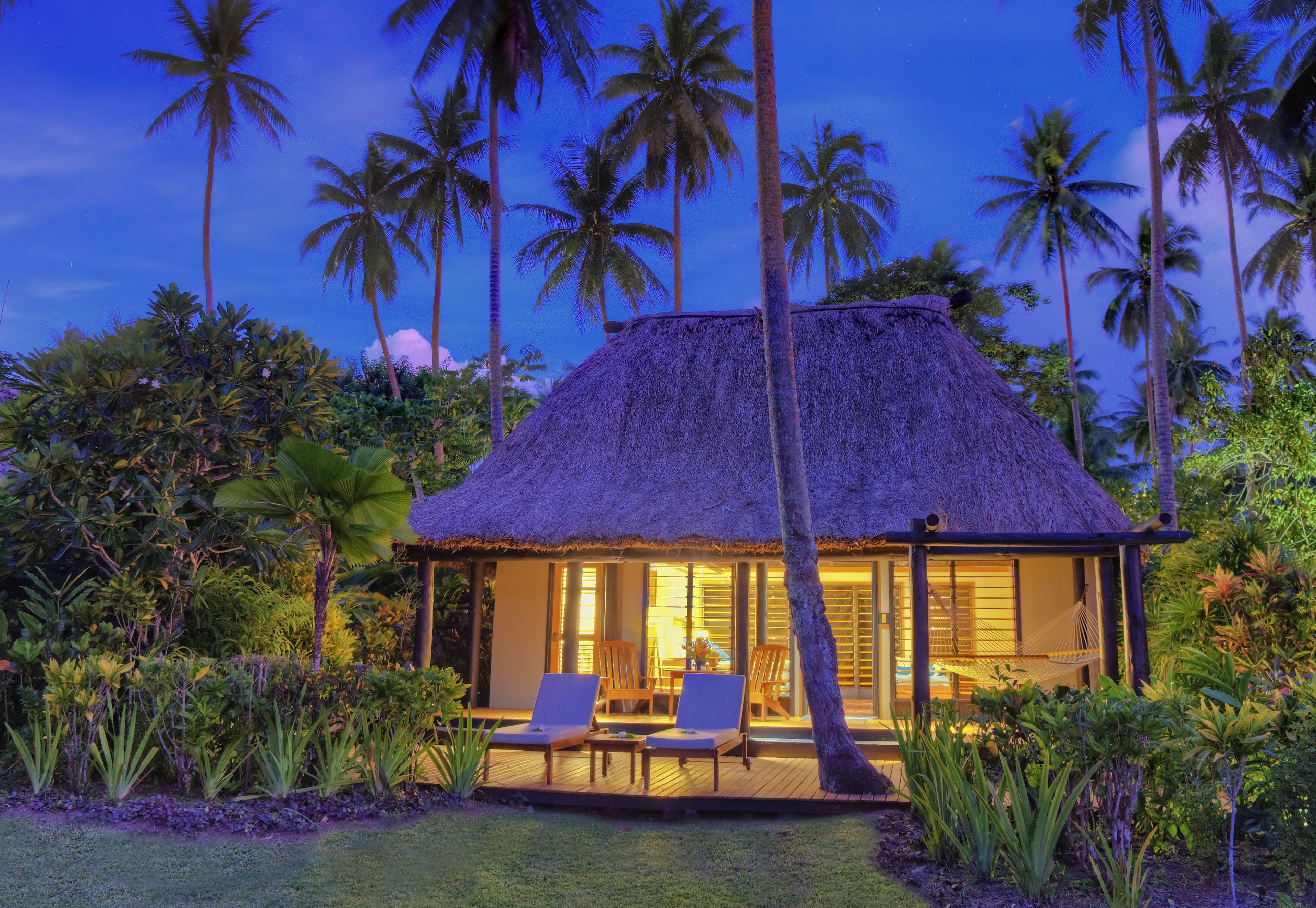 A thatched-roof bungalow with two lounge chairs and a dining table, surrounded by lush tropical vegetation.