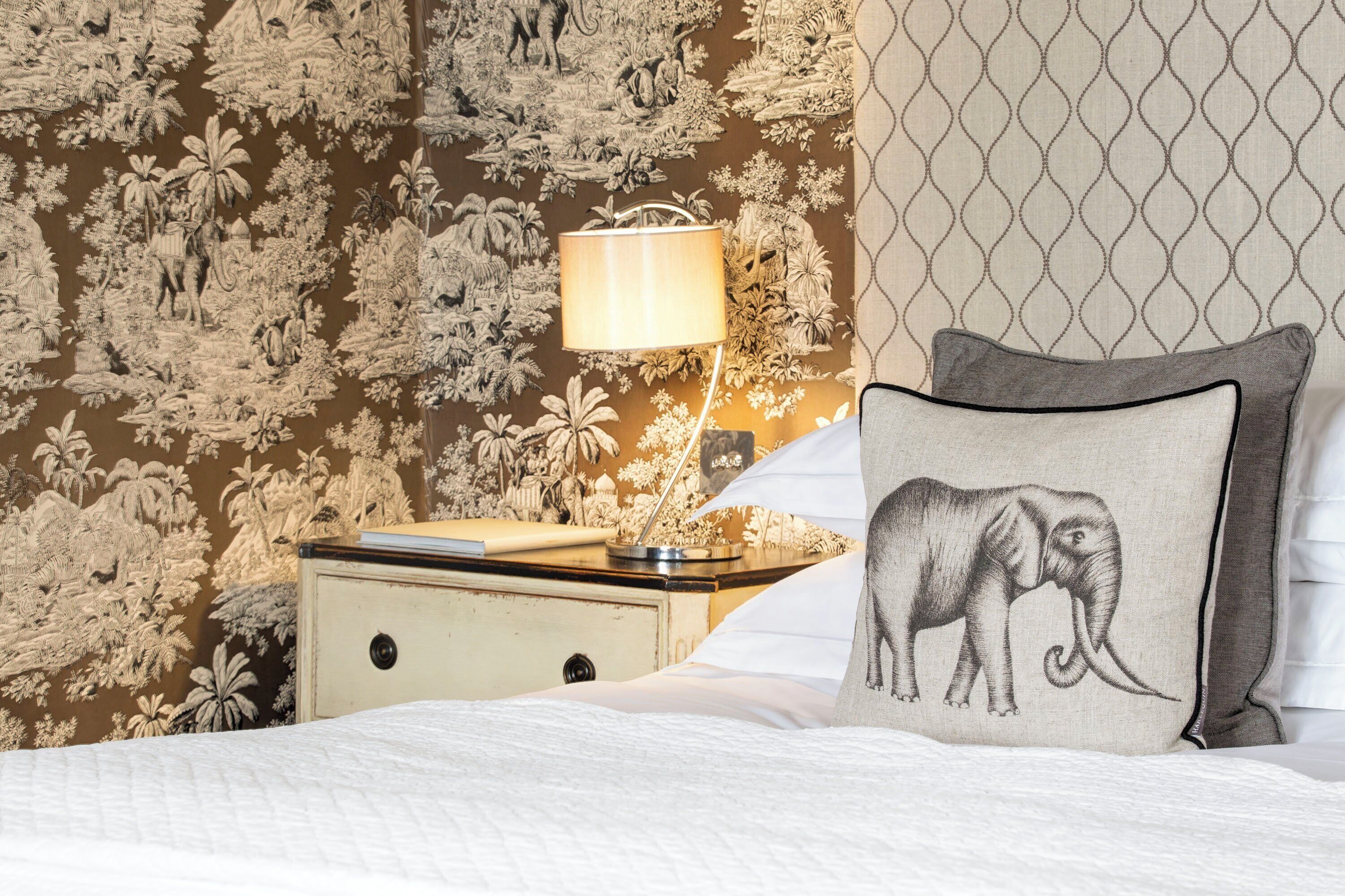 A neatly made bed with a pillow featuring an elephant design, a bedside lamp, and a nightstand with a book and a decorative item.
