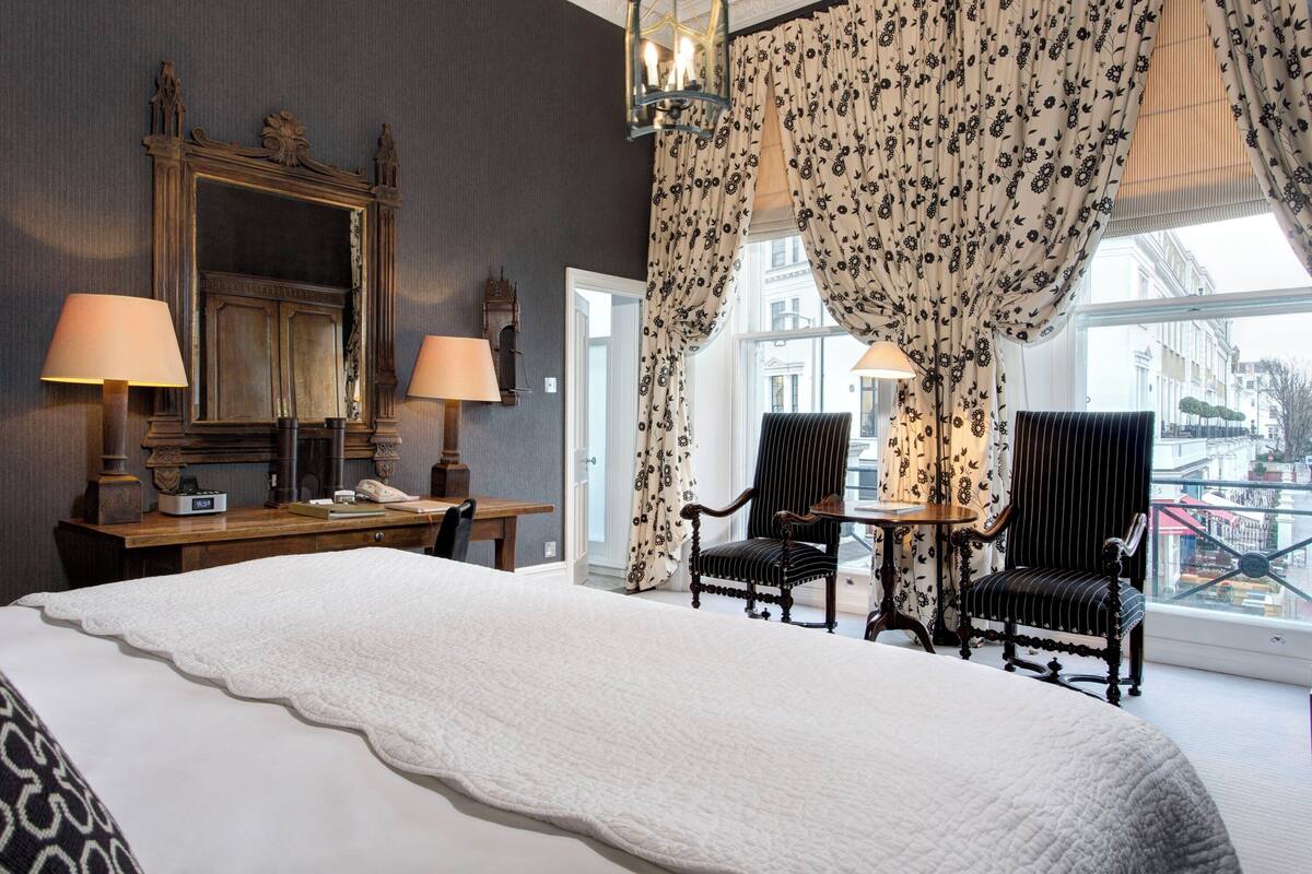 Habitación de hotel con una cama grande, dos sillas, un escritorio con lámpara, un espejo y vistas a la ciudad desde la ventana.