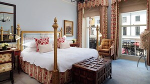 Una cama clásica con dosel, con cubrecama blanco y almohadas rojas y blancas, una mesita de noche de madera con una lámpara, una silla y una pequeña mesa con mantel de encaje.