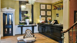 Un área de recepción con un escritorio de madera oscura, un jarrón blanco con flores, un sillón estampado y obras de arte enmarcadas en la pared.
