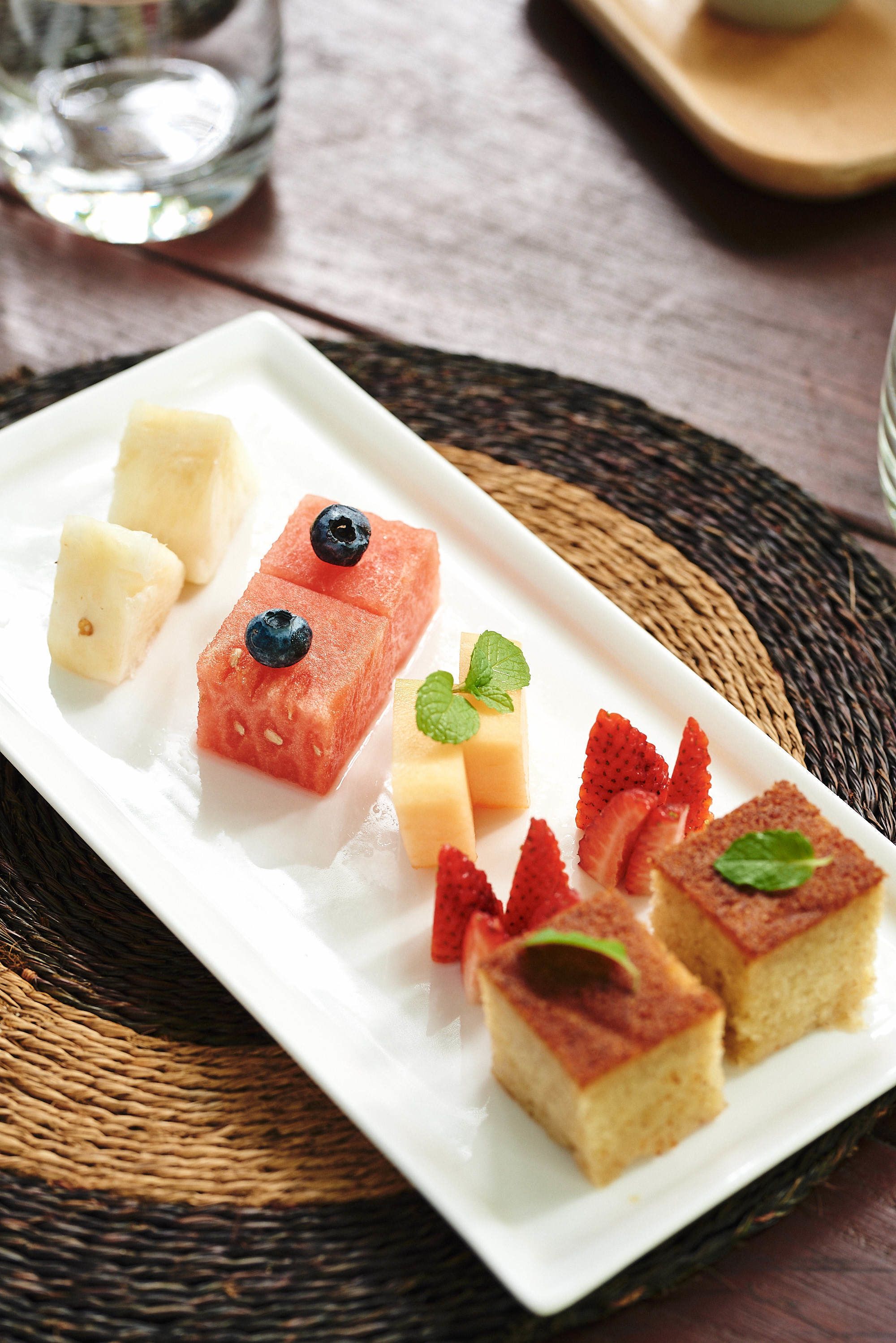 A white rectangular plate with various food items including watermelon, strawberries, and a cake, placed on a woven mat.