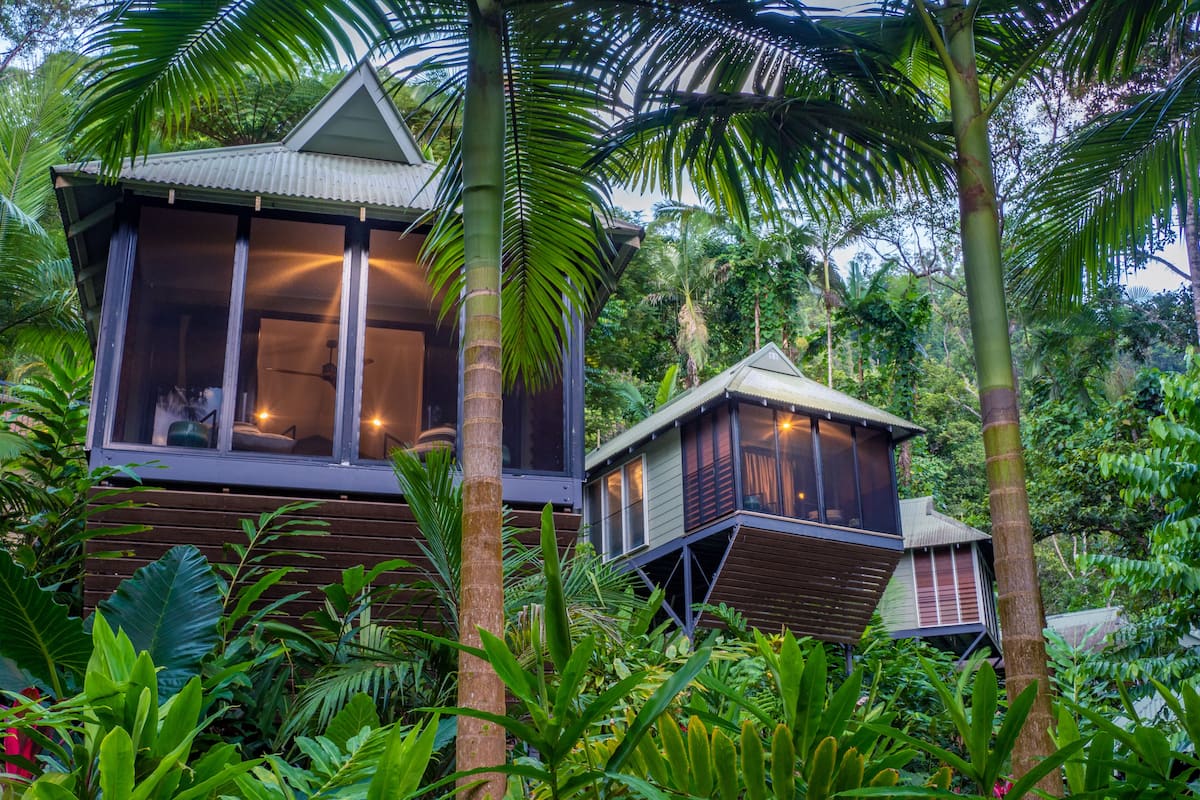 A wooden house with a large window and a balcony, surrounded by lush greenery.