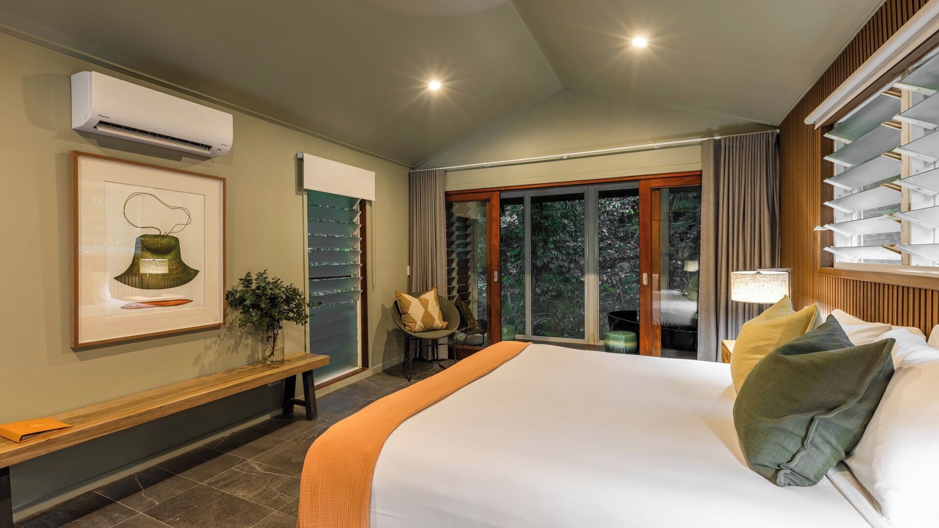 A hotel room with a large bed, a bench, a chair, a potted plant, and a view of the outdoors through a sliding door.