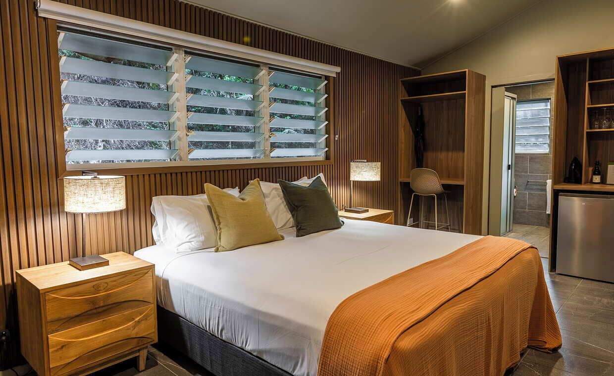 A modern bedroom with a large bed, wooden panel walls, a window with blinds, a nightstand, a desk, and a kitchenette area.