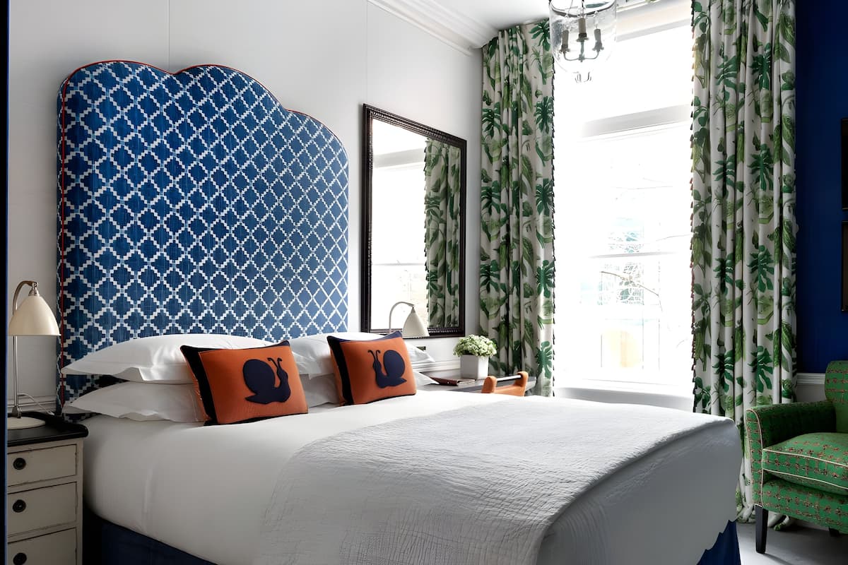 A bedroom with a large bed, two pillows with snail designs, a nightstand with a lamp, a dresser, a green chair, and a window with curtains.