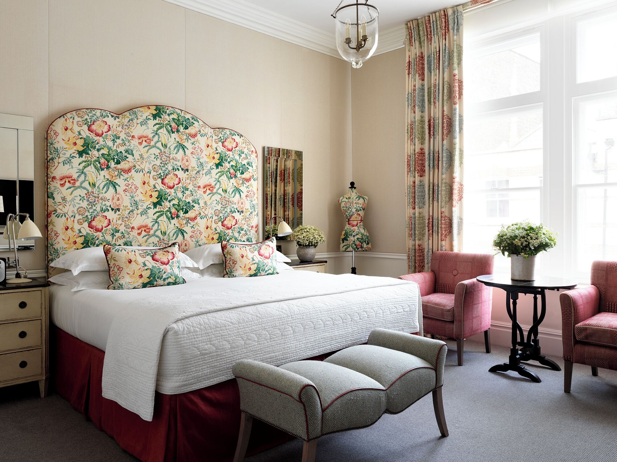 Quarto com uma grande cabeceira estampada com flores, uma cama com roupa de cama branca, uma mesinha de cabeceira com abajur, uma cadeira e uma pequena mesa com uma planta.