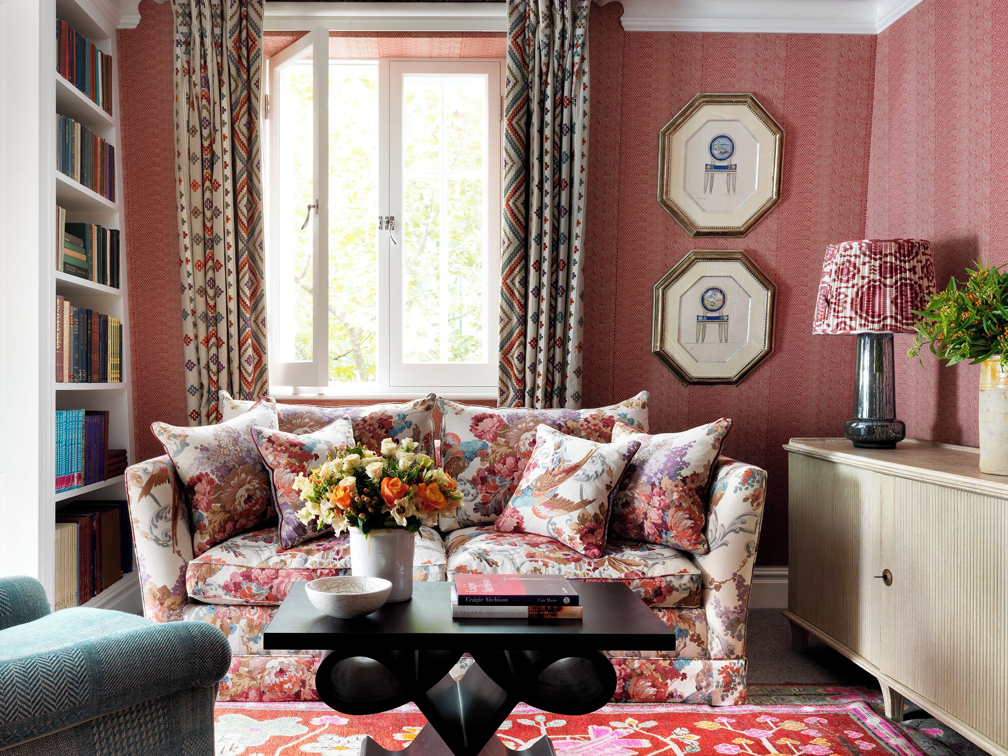 A living room with a sofa, a coffee table with a vase of flowers, a bookshelf, and a window with curtains.