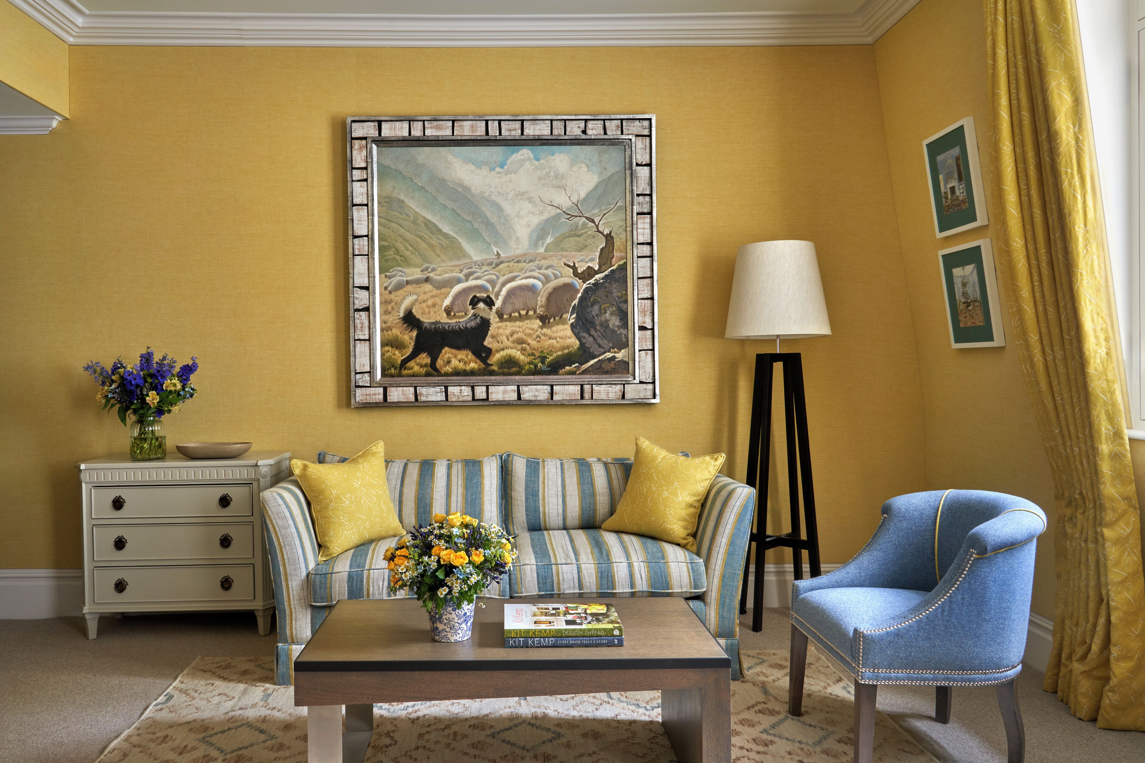 A room with a sofa, a chair, a coffee table, a lamp, a painting, and a vase with flowers.