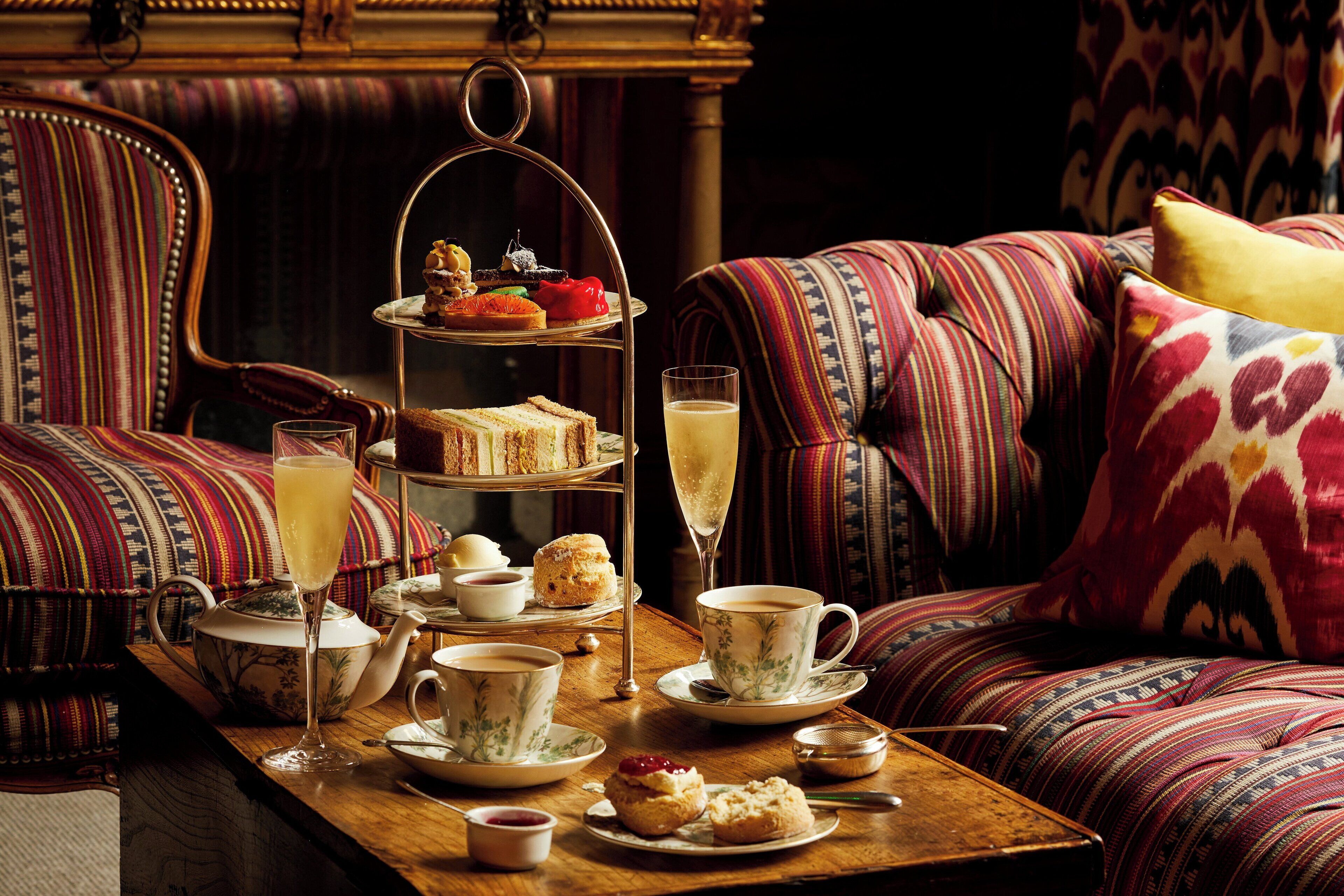 A cozy room with a wooden table set for afternoon tea, featuring a tiered tray with pastries, a teapot, cups, and a glass of champagne.