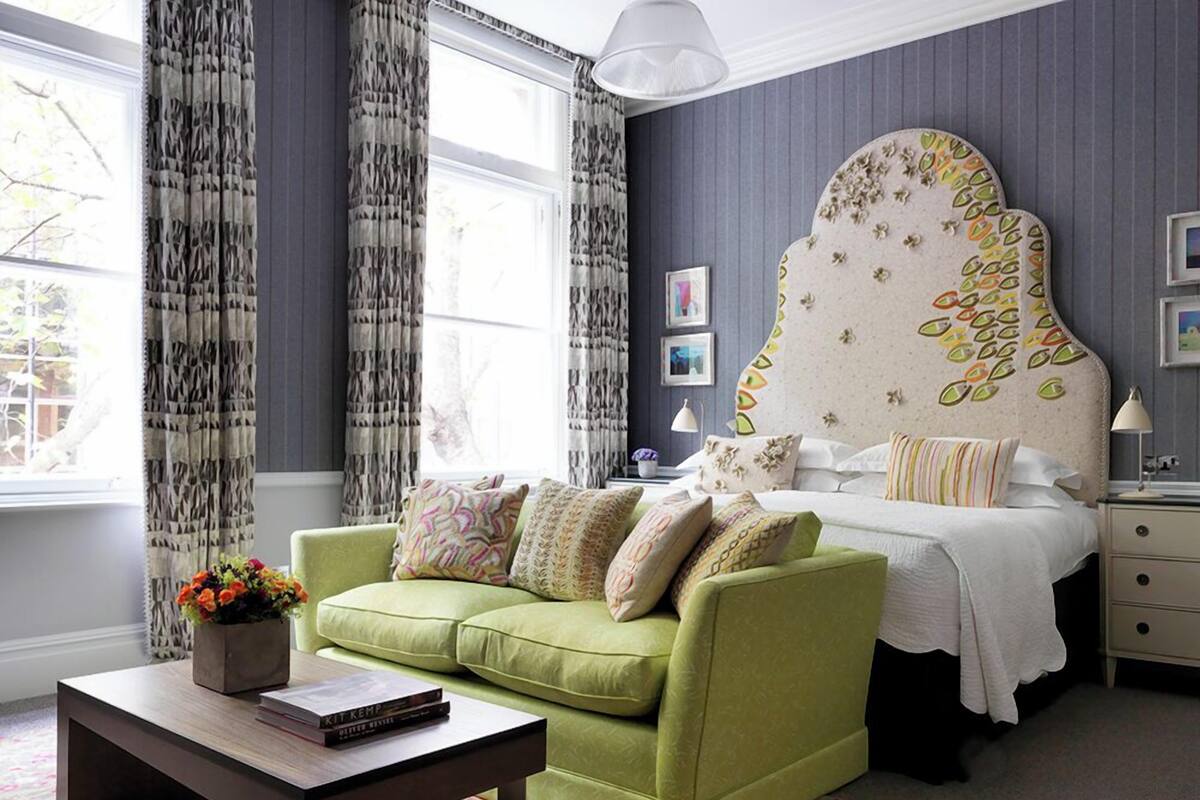 A bedroom with a green sofa, a bed with a decorative headboard, a bedside table, and a window with curtains.