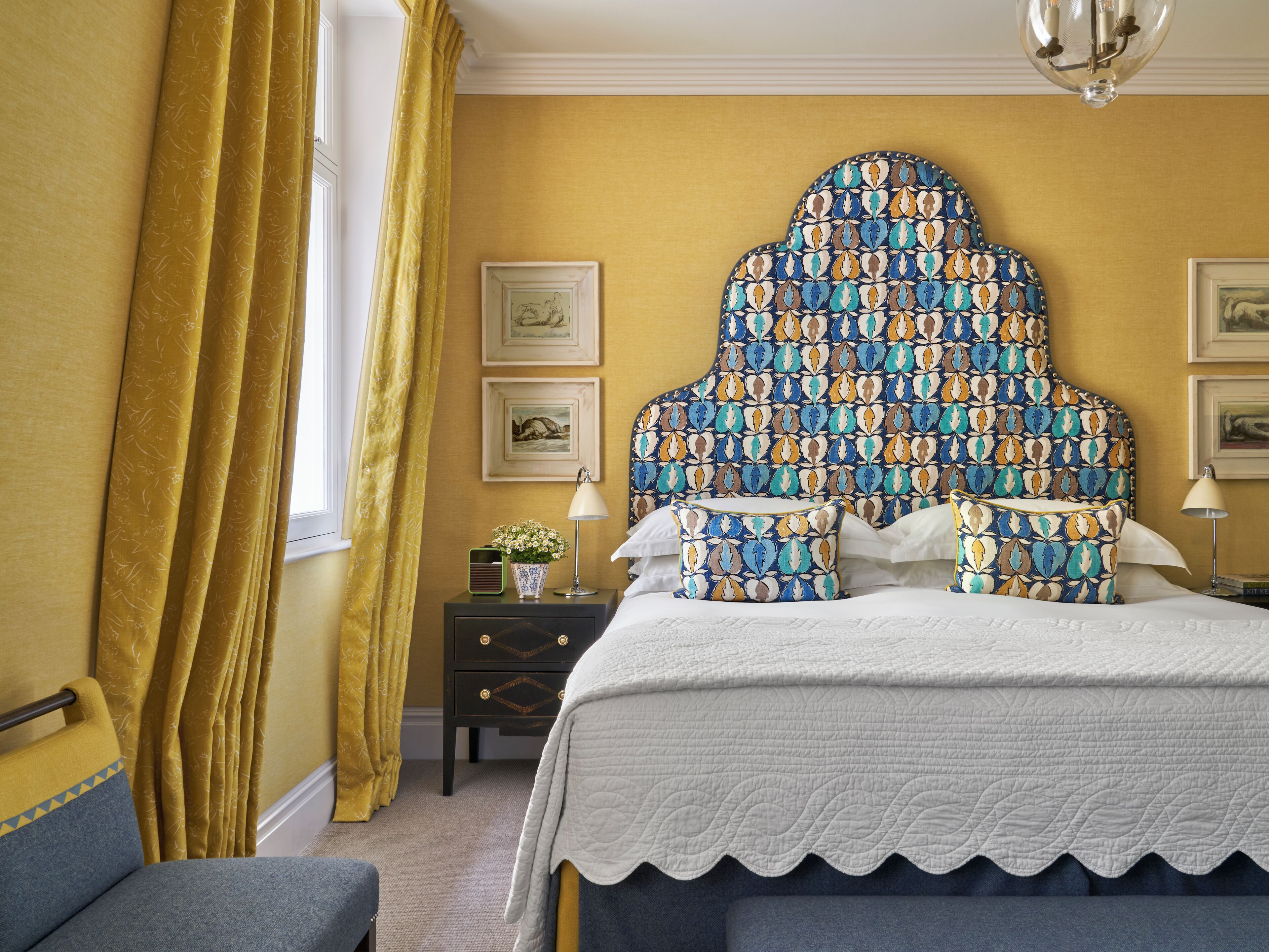 A bedroom with a patterned headboard, a bedside table with a lamp, and a window with curtains.