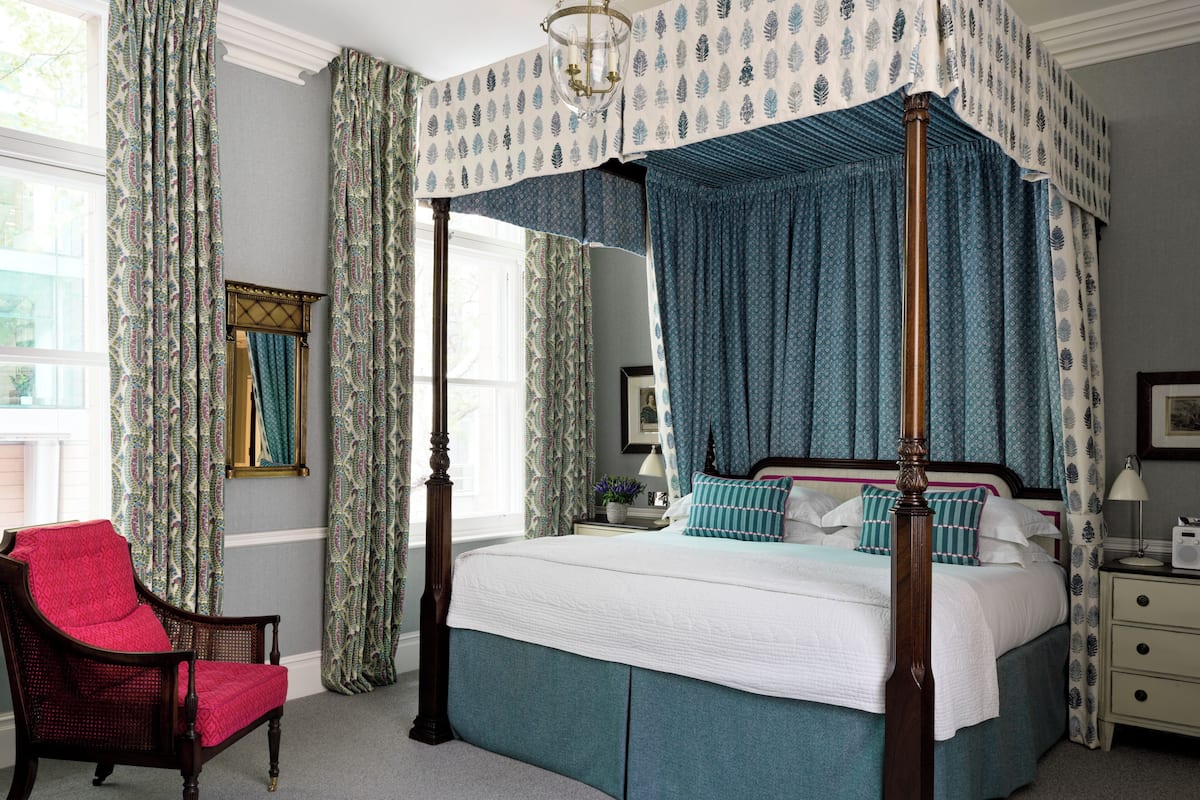 A four-poster bed with a canopy, a chair with a red cushion, and a bedside table with a lamp.