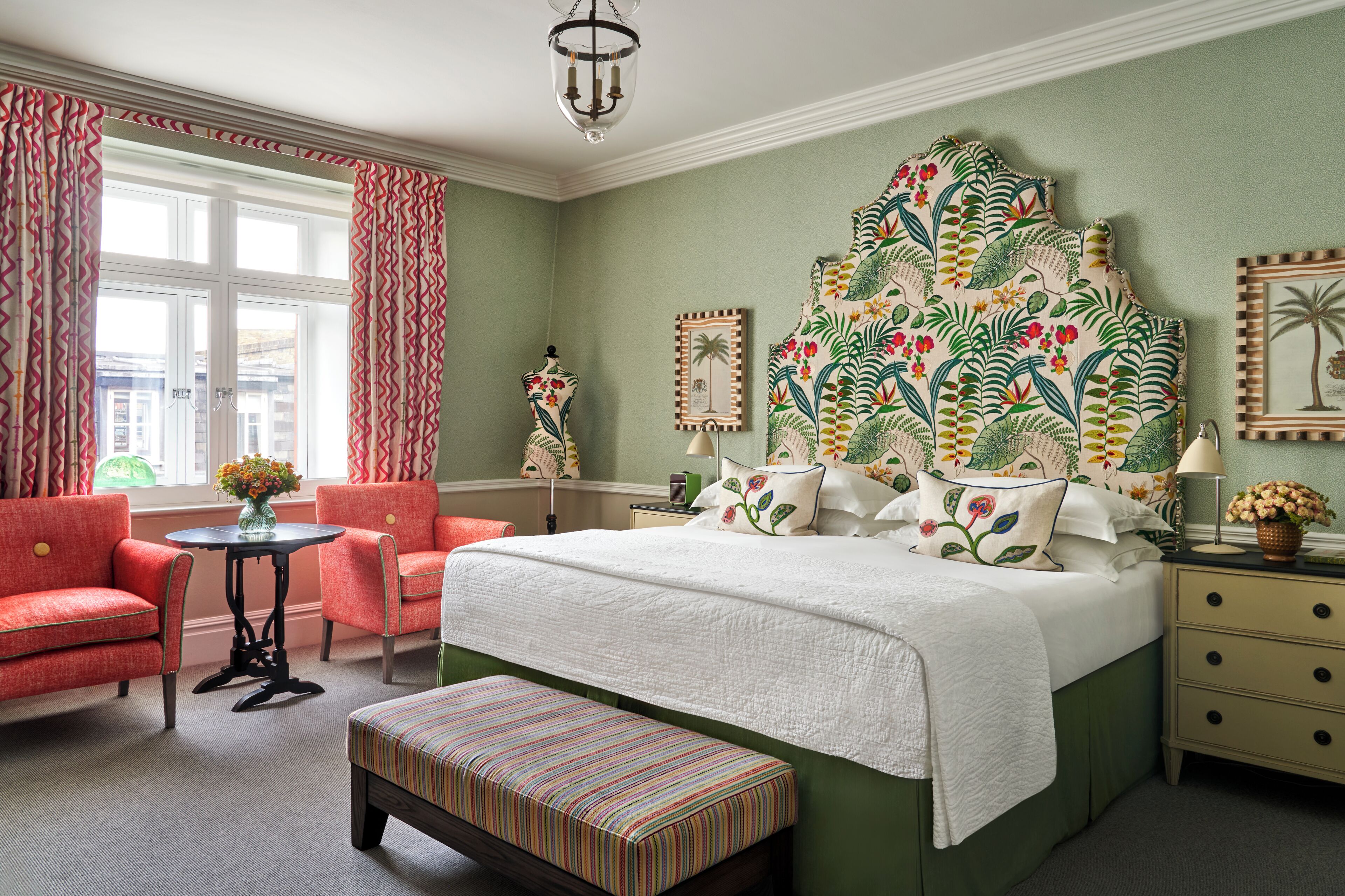 A bedroom with a large bed, a round table, two armchairs, a bench, and a nightstand with a lamp.