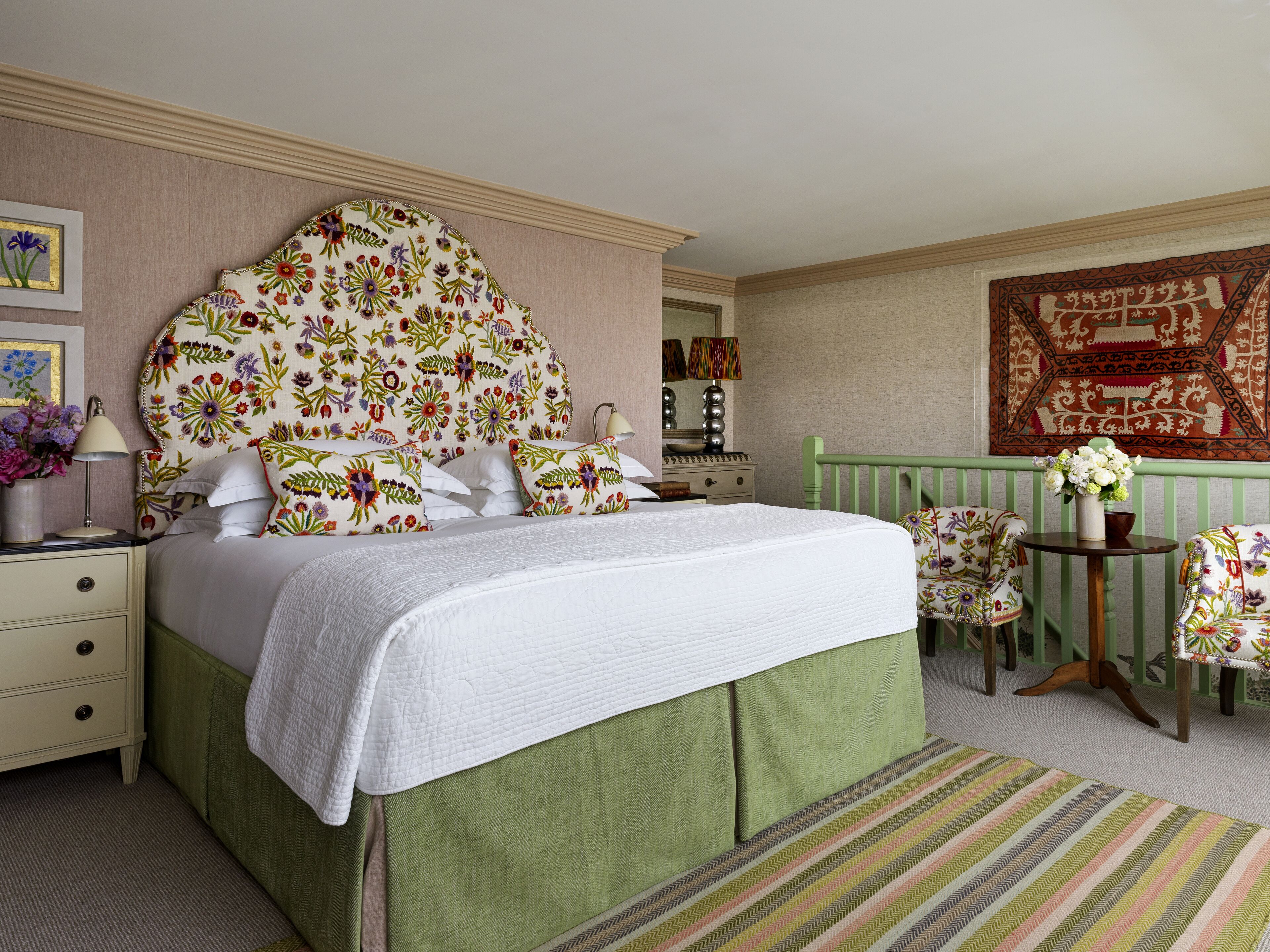 Quarto bem organizado com uma cama grande, uma cadeira verde, uma mesinha com um vaso de flores e um espelho sobre a cômoda.