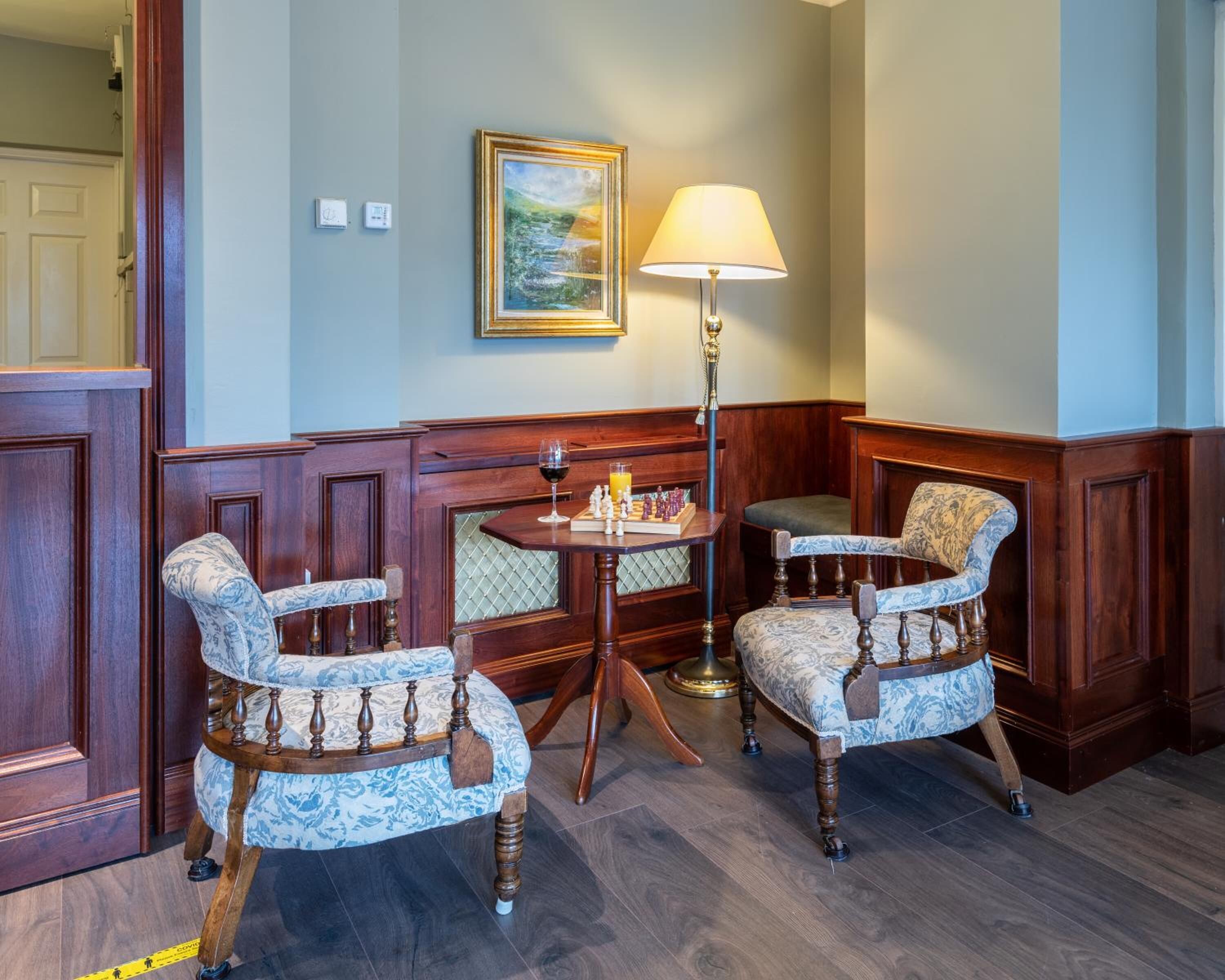 A corner seating area with two upholstered chairs, a small round table, a lamp, and a painting on the wall.