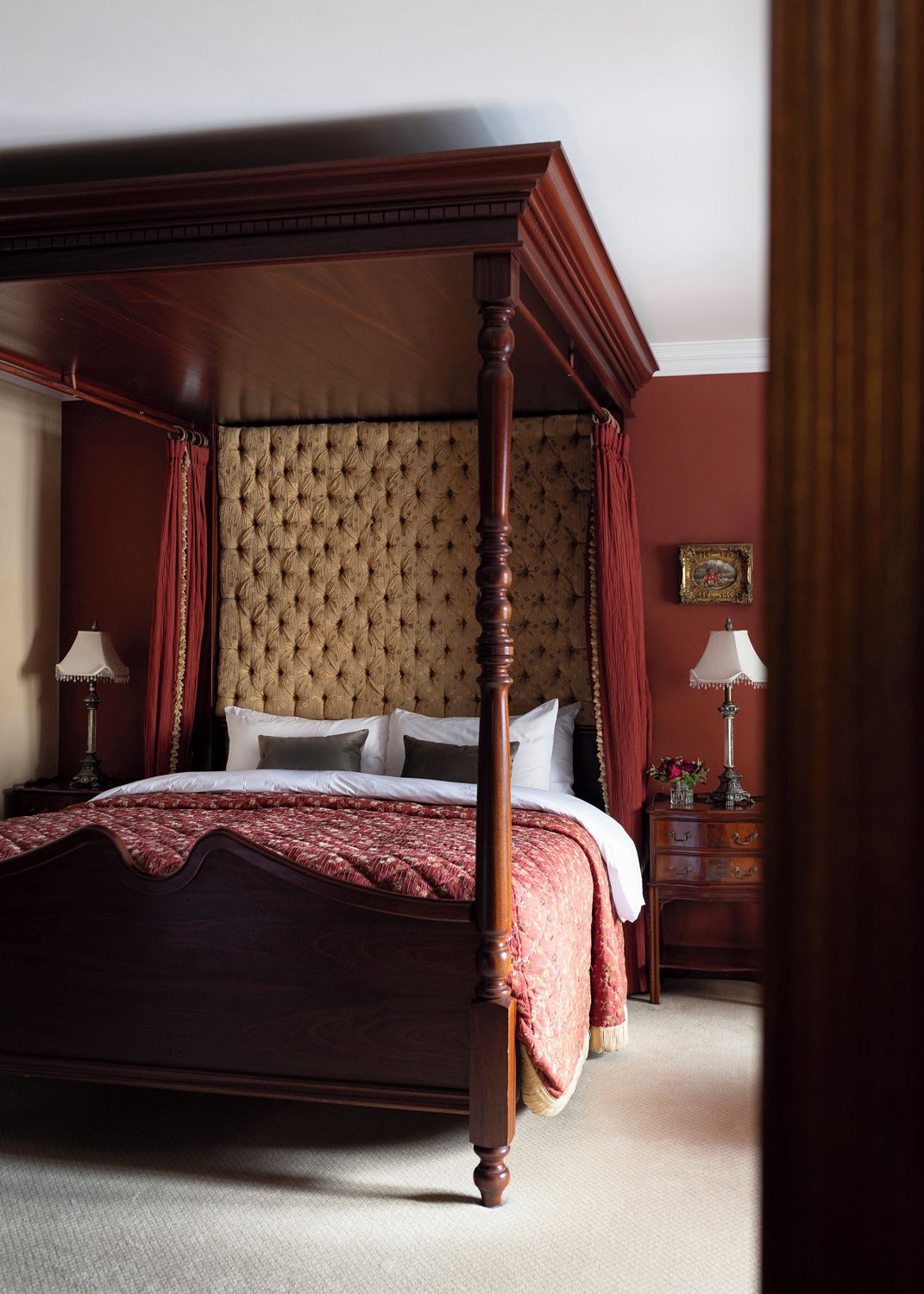 A four-poster bed with a tufted headboard, a bedside table with a lamp, and a nightstand with a lamp and a vase of flowers.