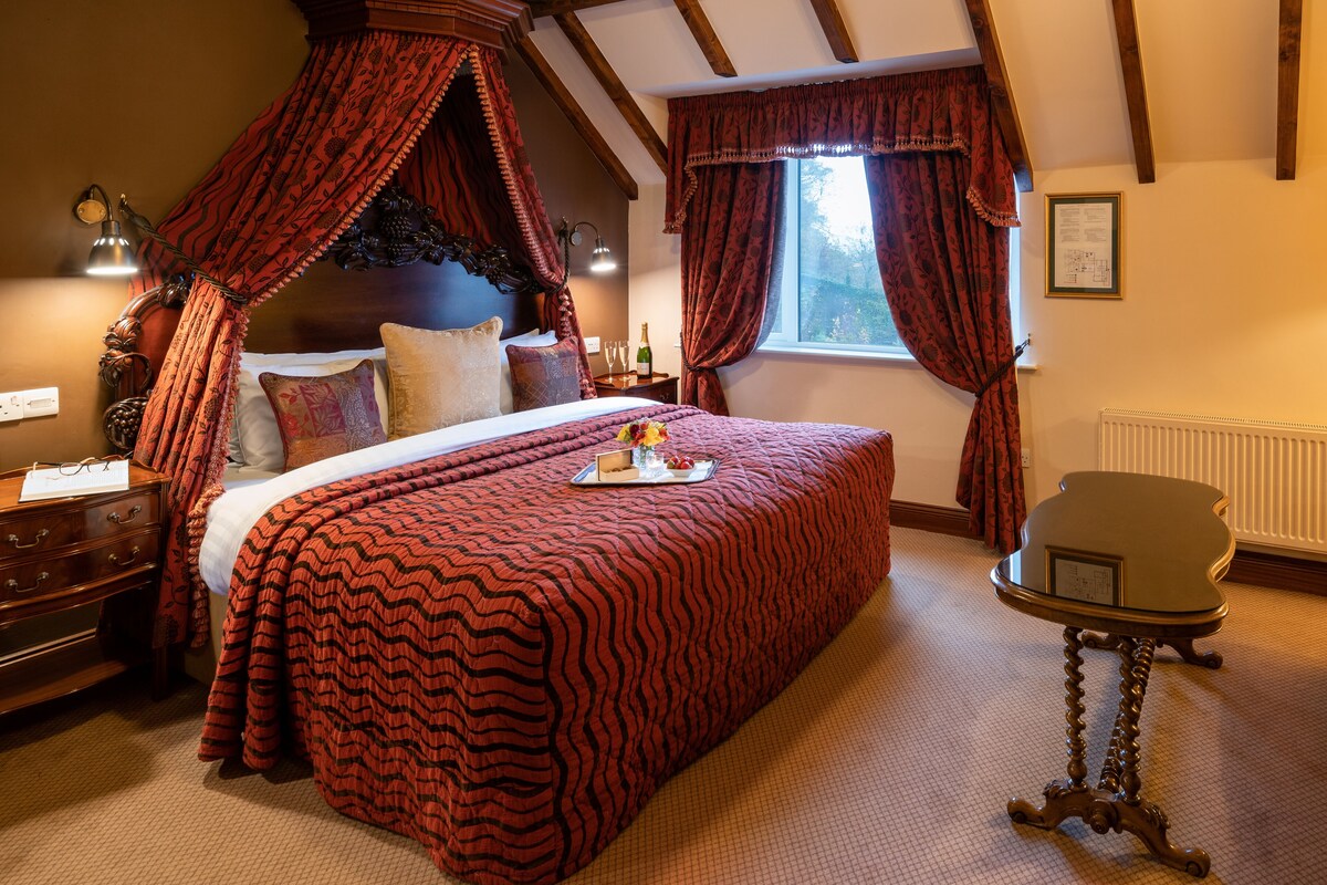 A bedroom with a four-poster bed, a nightstand, a bedside table, a window with curtains, and a radiator.