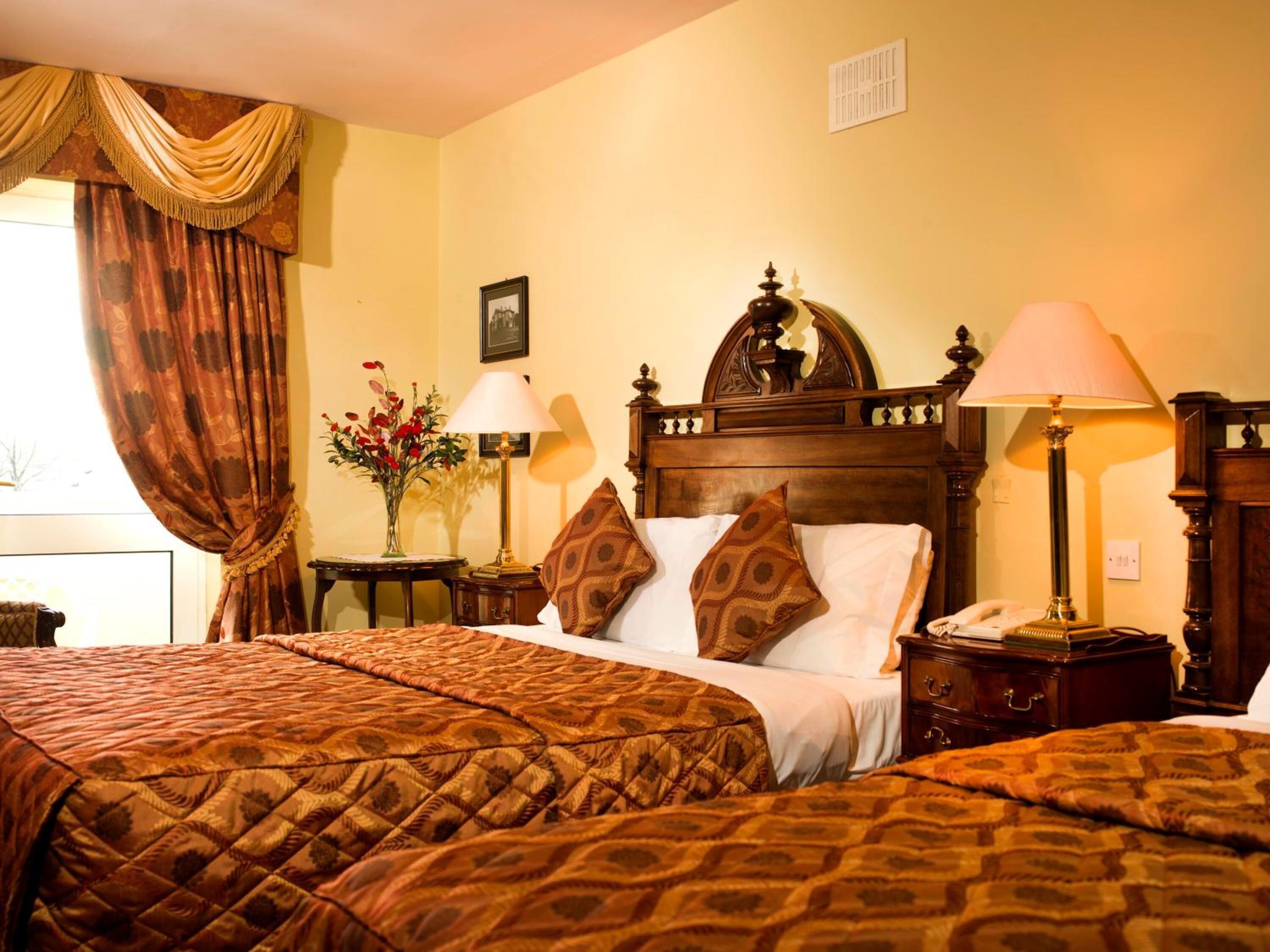 A hotel room with two beds, a wooden headboard, a nightstand with a lamp, and a vase with flowers.